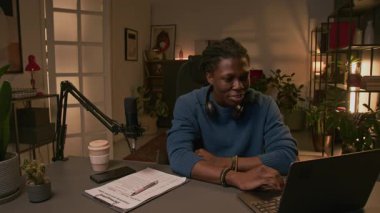 Orta halli Afrikalı Amerikalı erkek blogcu stüdyoda oturuyor, izleyicilere dizüstü bilgisayardan sorular okuyor, canlı talk show sırasında çevrimiçi izleyicilerle konuşurken cevap veriyor.