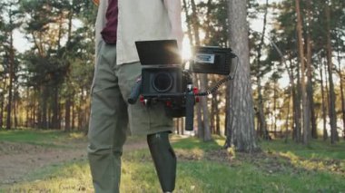 Sıcak güneşli bir günde elinde profesyonel video kamerayla ayakta duran, gündelik kıyafetli, bacak protezli, tanınmayan bir adamın orta kısmı.