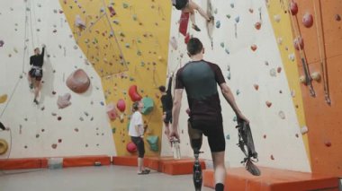 Protez bacaklı atletik adamın güvenlik ekipmanları ve su şişesiyle spor merkezinde duvara tırmanırken, bankta otururken ve protezi çıkarırken yavaş çekim.