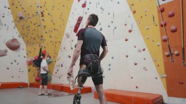 Arka görüş düşük açılı, bacak protezli erkek sporcunun orta derece yavaşlığı modern kapalı alan spor kompleksinde insanların duvara tırmanmasını izliyor.