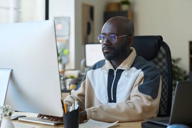 Gözlüklü ve günlük kıyafetli genç ciddi bir erkek tasarımcı bilgisayar ekranına bakıyor ve tuş takımının tuşlarına basıyor.