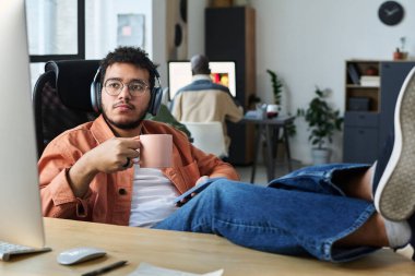 Dinlenmiş, kulaklıklı genç bir ofis çalışanı molada kahve içiyor ve özel ders izlerken bilgisayar ekranına bakıyor.