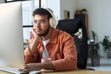 Kulaklıklı, bilgisayar ekranına bakan ve online video izlerken elini tuş takımına koyan genç, ciddi bir adam.