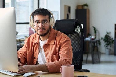 Gözlüklü ve kulaklıklı genç adam ağda kameraya bakıyor ve bilgisayar tuşlarına basıyor.