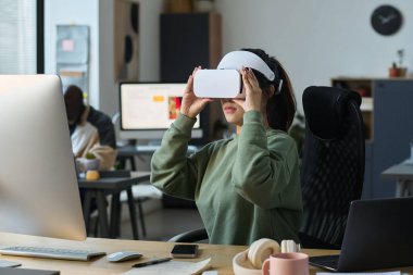 VR kasklı genç bayan tasarımcı işyerinde oturmuş proje üzerinde çalışırken sanal slaytlara bakıyor.
