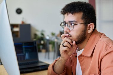 Genç düşünceli erkek programcı elini ağzında tutuyor ve bilgiyi çözerken bilgisayar ekranına bakıyor.