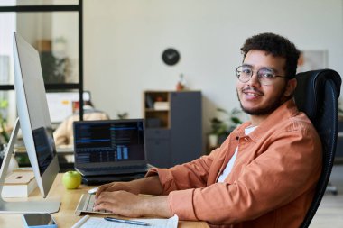 Genç gülümseyen erkek tasarımcı veya programcı yeni ürünü masaüstü bilgisayarı önünde test ediyor ve kameraya bakıyor