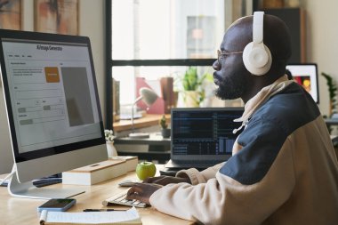 Kulaklıklı genç Afrikalı Amerikalı erkek tasarımcının ofiste görsel dil modeliyle bilgisayar ekranına bakışının yan görüntüsü