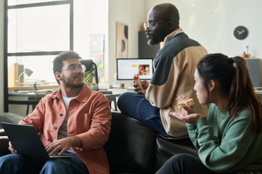 İki genç programcı öğle yemeği molasında sandviç yerken, bir tanesi bilgisayarlı erkek meslektaşıyla sohbet ediyor.