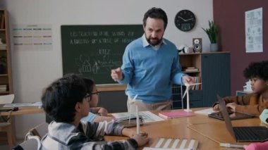 Çevresel farkındalık dersi sırasında bir grup çok ırklı genç öğrenciye rüzgar enerjisinden bahseden olgun erkek ekoloji öğretmeninin orta boy fotoğrafı. Güneş pili ve rüzgar türbini modelleri masalarda.