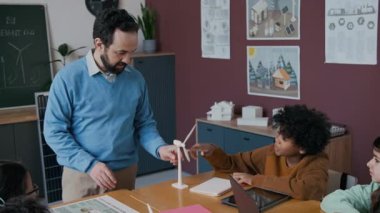 Orta yaşlı beyaz erkek öğretmen rüzgar enerjisi kavramını açıklarken ve çeşitli sınıflardaki çocuklara türbin modeli gösterirken çekilmiş orta yaşlı bir fotoğraf. Afrikalı Amerikalı çocuğun sorularını yanıtlıyor.