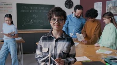 Orta sınıf inek bir orta sınıf öğrencisi gözlüklü, ekoloji dersi sırasında sınıfta dikilip kameraya bakıyor ve gülümsüyor, sınıf arkadaşları ve öğretmen tablete bakıyor.