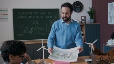 Çevre koruma posterlerini çocuklara göstererek, sonra kameraya bakıp gülümseyen, ekoloji dersi sırasında sınıfta duran neşeli, olgun, beyaz erkek fen öğretmeninin orta boy portresi.