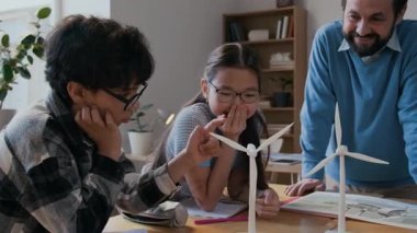 Orta boy Çinli kız ve Orta Doğulu çocuğun neşeli erkek fen öğretmeniyle rüzgar türbini modelleri üzerinde çalışırken, çalışma prensibini tartışırken, pervaneleri çevirirken ve sürdürülebilir teknolojiyi takdir ederken.