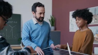 Yetişkin beyaz erkek öğretmenin, çevresel ders sırasında batarya ve fotovoltaik elementleri birlikte incelerken Afrika kökenli Amerikalı öğrencilerle güneş enerjisi teknolojisi hakkında konuşurken orta boy fotoğrafı.