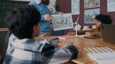 Tişörtlü ve gözlüklü genç bir öğrencinin ekoloji dersinde rüzgar türbinleri hakkında soru sormak için el kaldırdığı orta kavisli bir fotoğraf. Dinleyen ve cevap veren gülümseyen öğretmen.