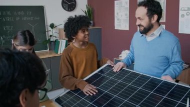 Çeşitli okul çocuklarına güneş enerjisi teknolojisini açıklayan, fotovoltaik hücreli pilleri ve siyah yüzeye dokunuşunu gösteren, bilgili, olgun beyaz erkek öğretmenin orta boy fotoğrafı.