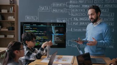 Orta yaşlı beyaz erkek kodlama öğretmenini dijital tabletle meşgul edip çeşitli okul çocuklarına yeni sorunları açıklarken orta ölçekli bir çekim, sonra da multimedya ekranındaki algoritma yapısına işaret ediyor.