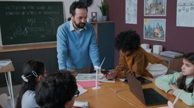 Orta yaşlı, neşeli beyaz erkek ekoloji öğretmeni çevresel eğitim dersleri sırasında çeşitli öğrencilere küçük taşınabilir güneş pili gösteriyor.