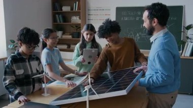 Orta yaşlı erkek öğretmenin ekoloji dersinde, sürdürülebilir elektrik üretim teknolojileri hakkında konuşan meraklı çok ırklı genç öğrencilerden oluşan bir gruba güneş paneli gösterirken orta yaşlı bir fotoğraf.
