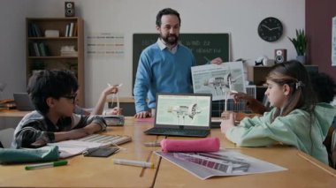 Orta boy Kafkas erkek fen öğretmeni yenilenebilir enerji kaynakları hakkında çok ırklı genç ilkokul öğrencileriyle konuşurken, dizüstü bilgisayar ekranında rüzgar türbinini gösteriyor