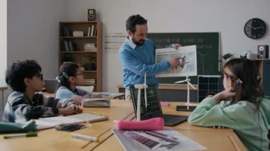 Orta boy erkek ekoloji öğretmeni dikkatli genç öğrencilere rüzgar türbini diyagramını gösteriyor, çalışma prensibini açıklıyor, sonra çocuk soru sormak için elini kaldırıyor.