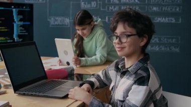 Orta ölçekli, orta ölçekli, gözlüklü bir Orta Doğulu öğrenci portresi. Kodlama dersinde masada oturuyor, dizüstü bilgisayarında algoritma üzerinde çalışıyor, kameraya bakıyor ve gülümsüyor.