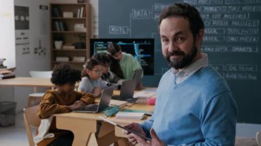 Orta yaşlı mutlu beyaz erkek kodlama öğretmeni, sakallı, tablet bilgisayarlı, sınıfta duran, kameraya bakan, gülümseyen ve dizüstü bilgisayarlarla çalışan öğrencilerin orta boy portresi.