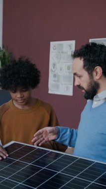 Kafkas erkek okul öğretmeninin 10 yaşındaki Afrikalı Amerikalı öğrenciye güneş enerjisi teknolojisini açıklarken sınıfta fotovoltaik elementlerin olduğu panele bakarken orta boy dikey görüntüsü.