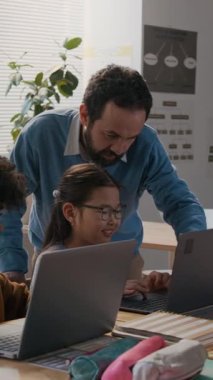 Orta boy beyaz erkek kodlama öğretmeninin ders sırasında sınıfta dolaşması, çeşitli öğrencilerin dizüstü bilgisayarlara program yazmasına yardım etmesi, hatalara ve zorluklara yardım etmesi,