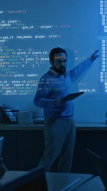 Erkek programlama öğretmeninin okul veya üniversite dersleri sırasında dizüstü bilgisayarlı öğrencilere yüzlerine semboller ve rakamlar yansıtılan karanlık sınıflarda yeni bilgisayar senaryosu açıklarken tam dikey görüntüsü.