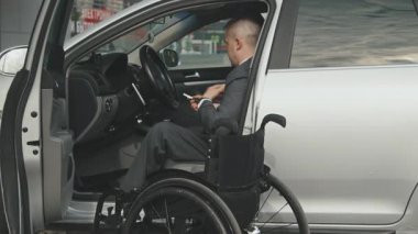 Sürücü koltuğunda oturan ve tekerlekli sandalyeyle arabadan inen işadamının el kamerasıyla çekilmiş görüntüsü.