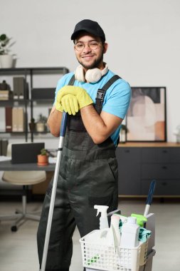 Elinde paspas, kovayla kameraya gülümseyen üniformalı temizlikçi portresi.