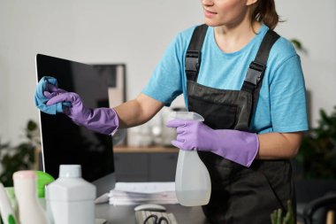 Ofisi temizlerken bilgisayar ekranını bez parçasıyla silen üniformalı temizlikçi kadın.