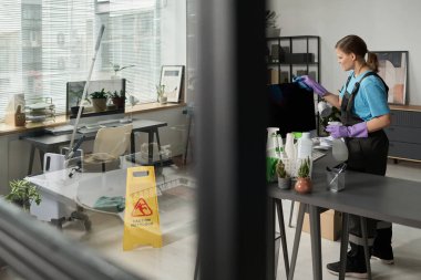 Temizlikçi üniformalı, temizlik sırasında bilgisayardaki tozu temizliyor.