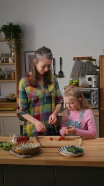 Tam dikey çekimde, beyaz kız annesine öğle yemeği için sağlıklı salata yaparken, domates soyarken ve gülümseyen anne sebzeleri dilimlerken sohbet ederken yardım ediyor.