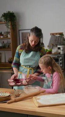 Orta boy dikey çekimde elinde kek tepsisi olan neşeli beyaz anne ve önlüklü genç kız, Pazar günü evde birlikte pişirirken ve pişirirken hamuru dikkatlice dolduruyorlar.