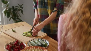 Kafkasyalı annenin mutfaktaki tahta tahtada salata için taze sebze keserken orta eğik pozu, sonra da masada oturan ve neşeyle gülümseyen küçük kıza bir parça salatalık verirken.