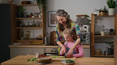 Orta boy gülümseyen olgun bir beyaz kadın ve 7 yaşında bir kız çocuğu mutfakta duruyor. Anne, kıza salata için sebze kesmeyi öğretiyor. Salatalıktan ısıran ve gülen bir kız.