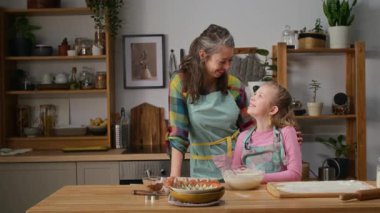 Mavi önlük giymiş neşeli beyaz bir annenin orta boy portresi ve evde duran 7 yaşındaki kız çocuğu kek pişirmek için hamur işi yapıyor, sonra kameraya bakıyor ve gülümsüyor.