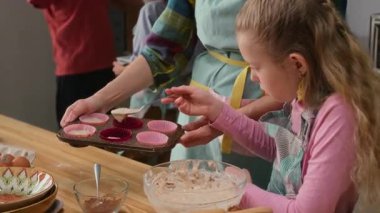 Orta boy sarışın beyaz önlüklü kız kek tepsisini dikkatlice hamurla doldururken annenle evde yemek pişirirken, kardeşi arka planda tabak getirirken.