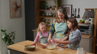 Küçük kız kardeşi izlerken orta boy beyaz bir çocuğun aile evindeki mutfakta kurabiye ya da kek pişirmek için annesine yardım ederken, un dökerken görüntüsü.