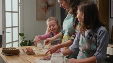 Annesi hamuru çırparken, annesi un döken iki beyaz kardeşin orta boy fotoğrafı. Hafta sonları turta pişirmeye hazırlanırken erkek çocuk un tozunu alıyor.