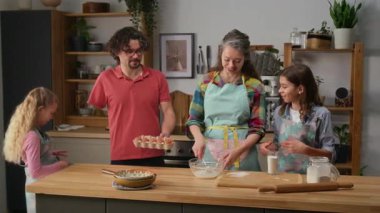 Orta ölçekli gülümseyen beyaz kadın mutfakta duruyor, kek için hamur işi çırpıyor, oğluyla ve kızıyla sohbet ediyor, ve uzuvları farklı bir şekilde uzuvlarıyla kocasını seviyor yumurta getiriyor ve karısını öpüyor.