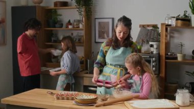 Orta boy gülümseyen beyaz anne ve genç kızı önlükleriyle kek tepsisine hamuru dökerken, engelli babası ve ergen oğlu da yemek tabakları getiriyor.