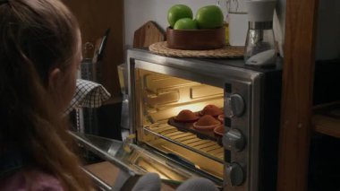 Kafkasyalı mutlu bir kadının sıcak fırından yeni çıkmış keklerle tepsiyi kaldırmak için yastıklı eldiven giymesine yardım ettiği, sonra hamur tatlısı kokusu ve neşeyle gülümsediği orta kavisli bir fotoğraf.