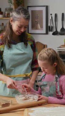 Orta boylu, olgun, beyaz bir annenin önlük giydiği, sarışın bir kızın kek hamuruna kakao kattığı ve hafta sonları veya tatillerde birlikte yemek pişirirken kakaoyu karıştırdığı dikey bir fotoğraf.