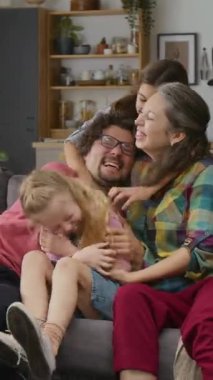 Orta dikey çekimde kolunda sakatlık olan beyaz bir baba, mutlu bir anne, genç bir oğlan ve genç bir kız kanepeye oturmuş, sarılıyor, gıdıklama oyunu oynuyor ve neşeyle gülüyor.