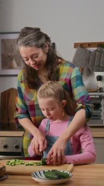 Mutfakta dikilen mutlu beyaz kadın ve genç kızın orta boy dikey pozu. Anne, öğle yemeğinde sağlıklı salata için kıza bıçak tutmayı ve salatalık ve domates doğramayı öğretiyor.