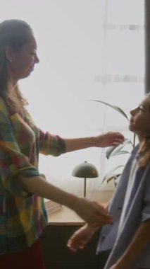 Beyaz anne ve genç oğlanın birbirine doğru adım attığı, sarılıp nazikçe öpüştüğü, oturma odasındaki parlak pencerenin önünde orta boy dikey bir çekim.